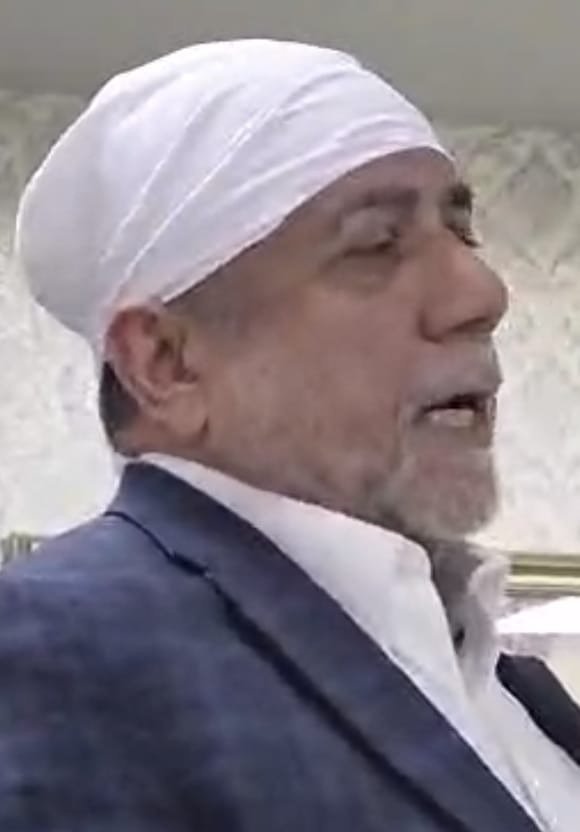 A man in a white head covering and dark jacket at the gathering.